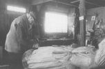 Original title:  Photographie d’archives en noir et blanc. Un homme sculpte le pied d’une grande statue en bois à l’intérieur d’un atelier où sont éparpillés croquis et outils servant à travailler le bois.